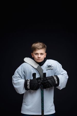 A young hockey player poses in a white jersey and black gloves against a dark background.