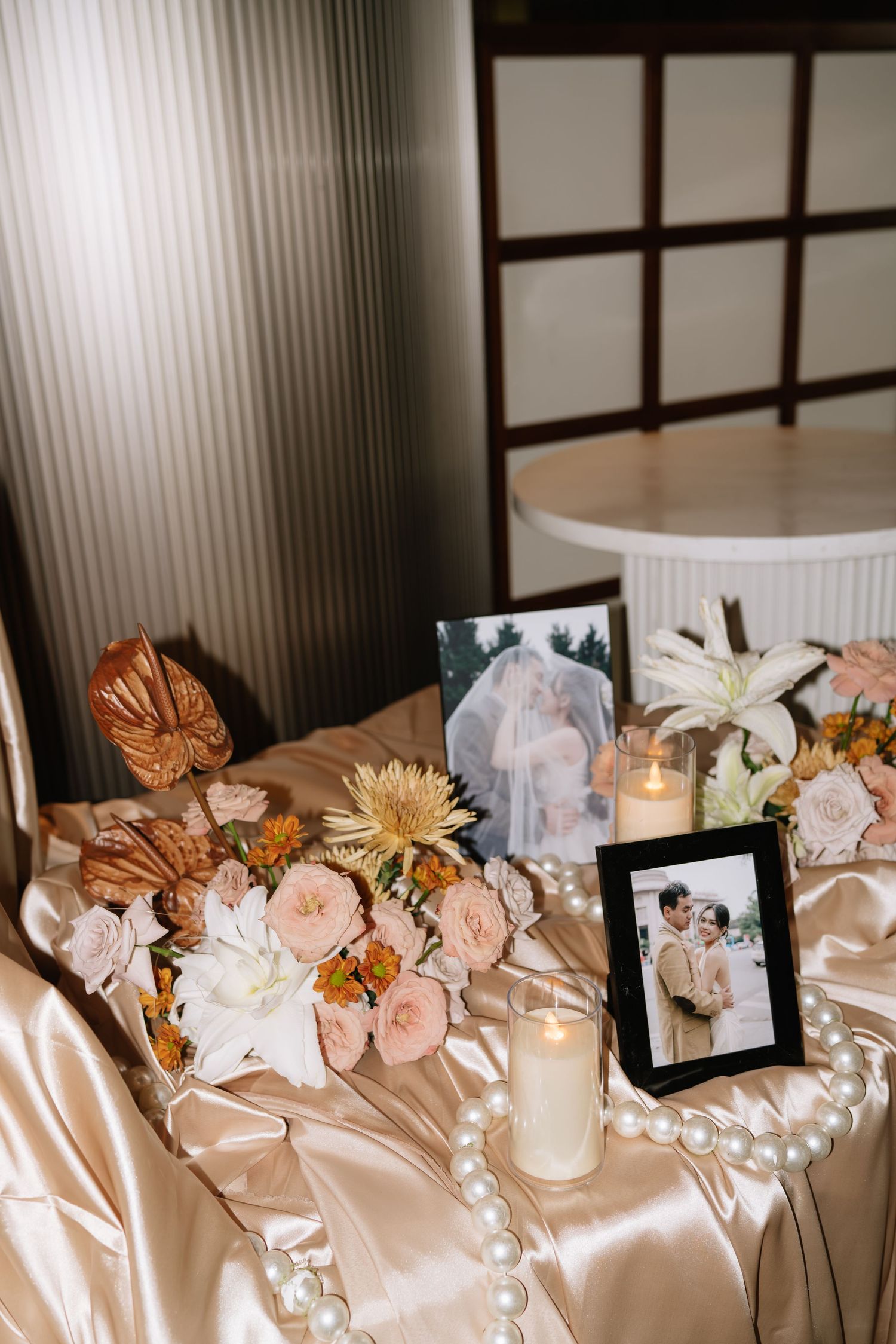 Wedding table display with framed couple photos, candles, pearls, and floral arrangements on satin cloth.