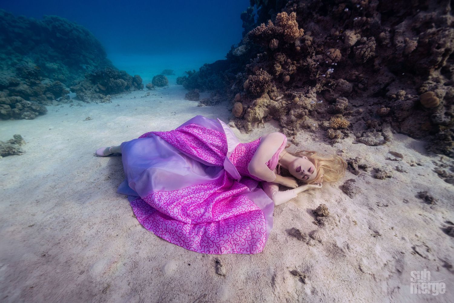 Flowing pink fabric creates elegant shapes underwater against a rocky ocean floor background.