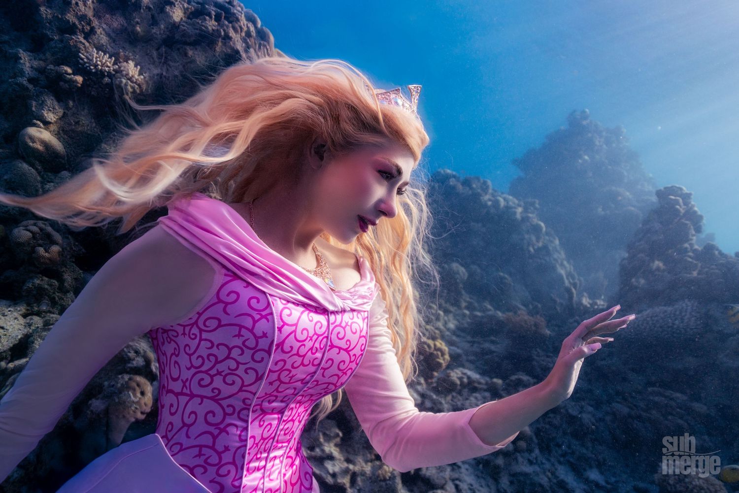 Pink dress flowing gracefully in crystal clear underwater scene with dramatic rock formations.