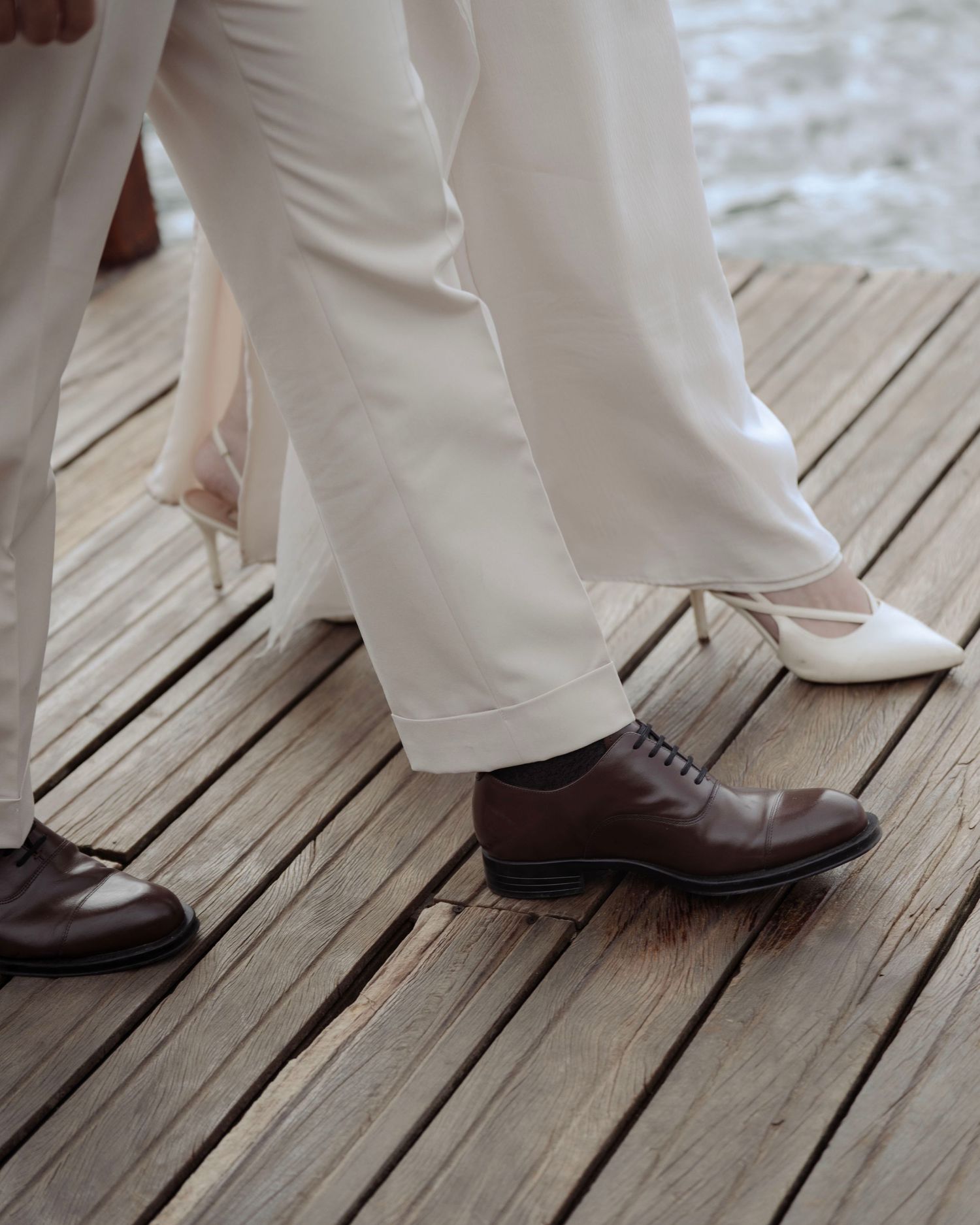 Close-up of bride in white heels and groom in brown oxford shoes on wooden deck.