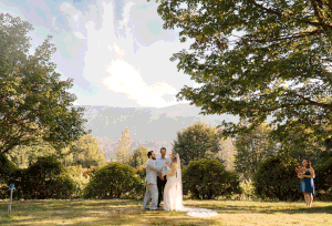 A couple in wedding attire poses in a scenic outdoor setting with mountain views and lush trees in the background.