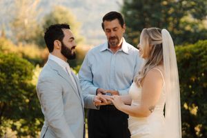 A couple shares an intimate moment during their outdoor wedding ceremony as sunlight filters through trees in the background.