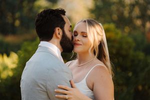 A couple embraces romantically in an outdoor wedding setting with glowing natural sunlight in the background.