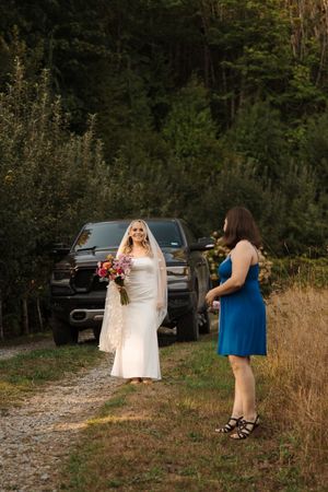 A bride in a white lace dress stands on a dirt path next to someone in blue, with a black truck in the background.