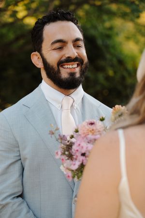 A bearded person in a light gray suit and pink tie smiles joyfully during an outdoor wedding ceremony.