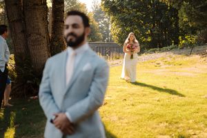 A bride in white dress approaches from behind as groom in light blue suit stands in sunny garden setting during first look.