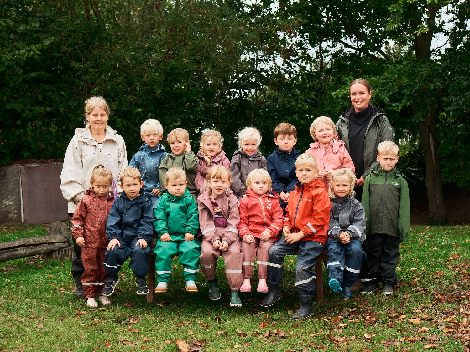 A group of young children sit together outdoors wearing colorful jackets on a wooden bench in autumn.