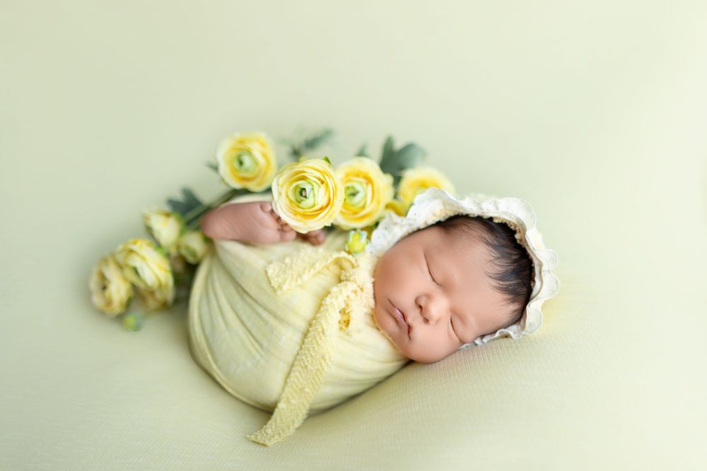A sleeping baby in a yellow outfit lies peacefully surrounded by soft yellow rose flowers.