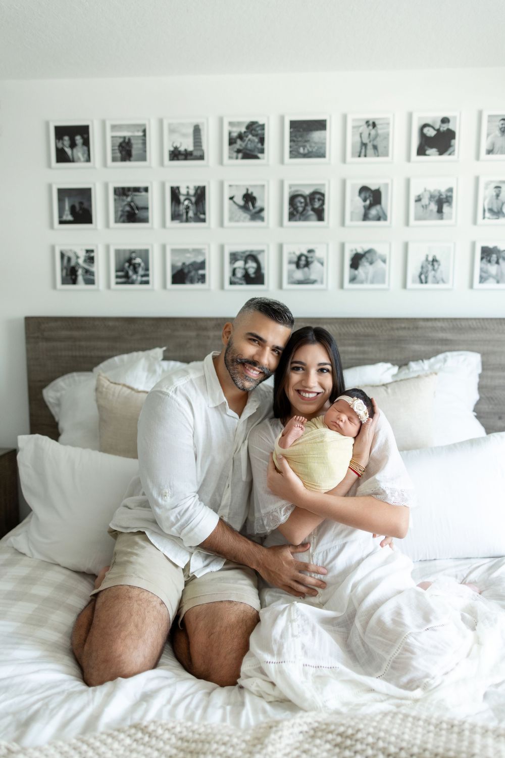Three people relaxing together on a cozy white bed with framed photos displayed on the wall behind them.