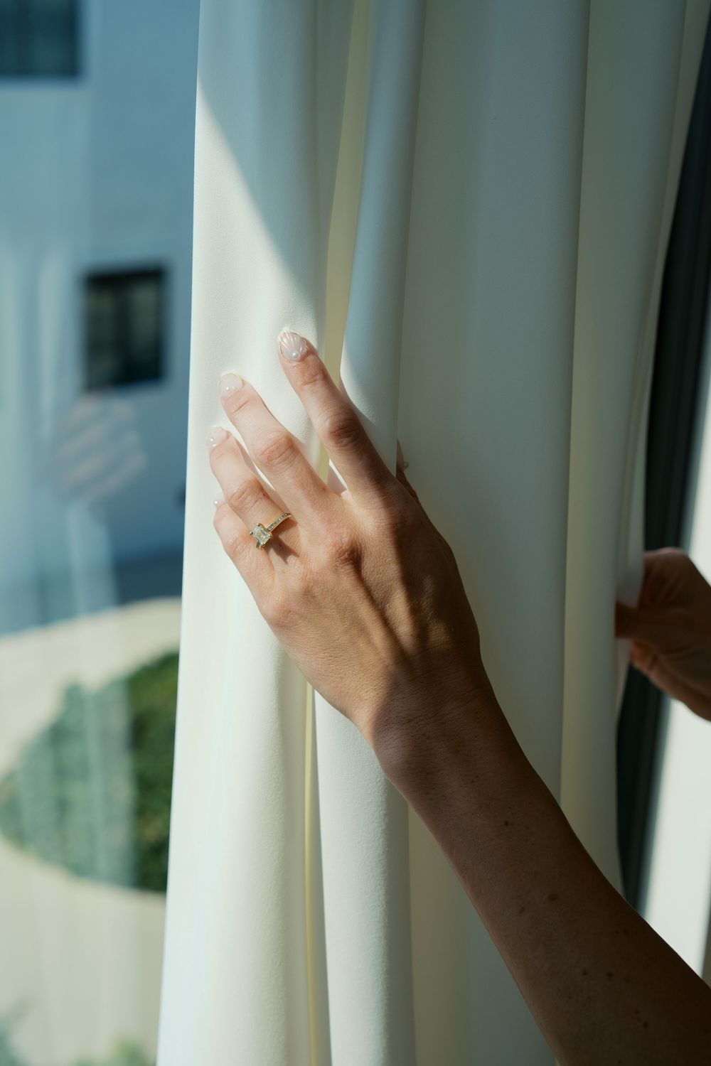 A delicate hand wearing an engagement ring brushes against white curtains in soft window light.