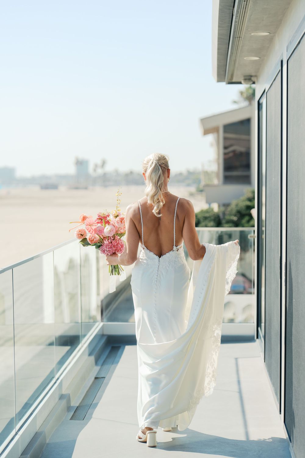A figure in a white dress views the cityscape from a balcony with pink flowers in golden sunlight.