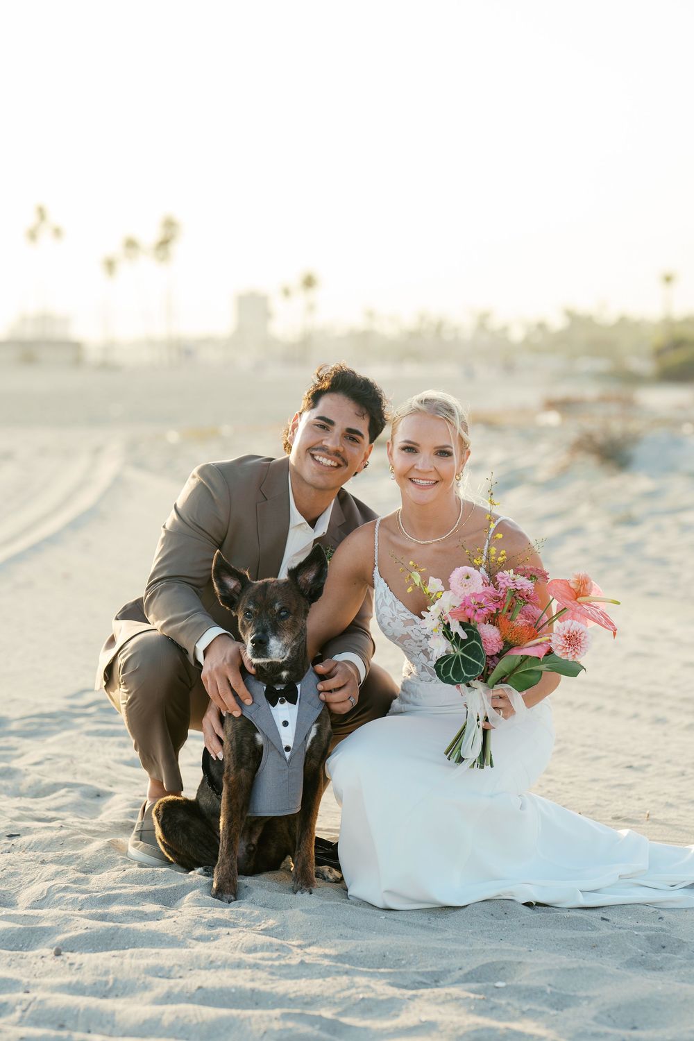 Wedding couple sits on beach with bouquet of tropical flowers at sunset.