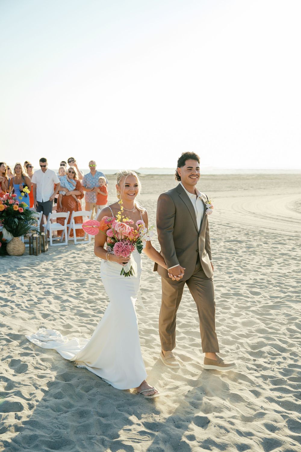 Beach wedding ceremony procession with guests watching the celebration on white sand.