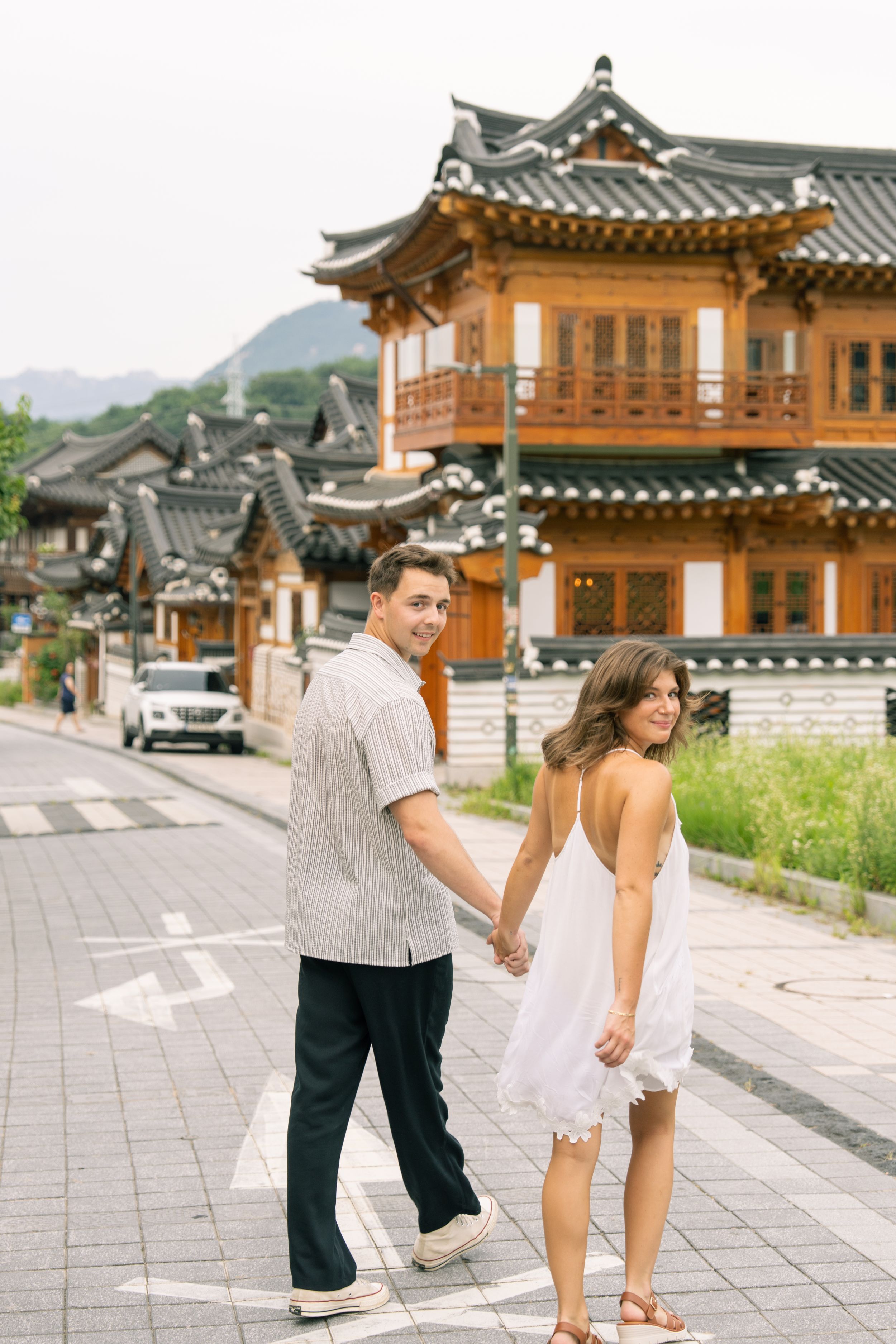 Couple enjoying a playful moment in front of traditional Korean buildings and mountains.