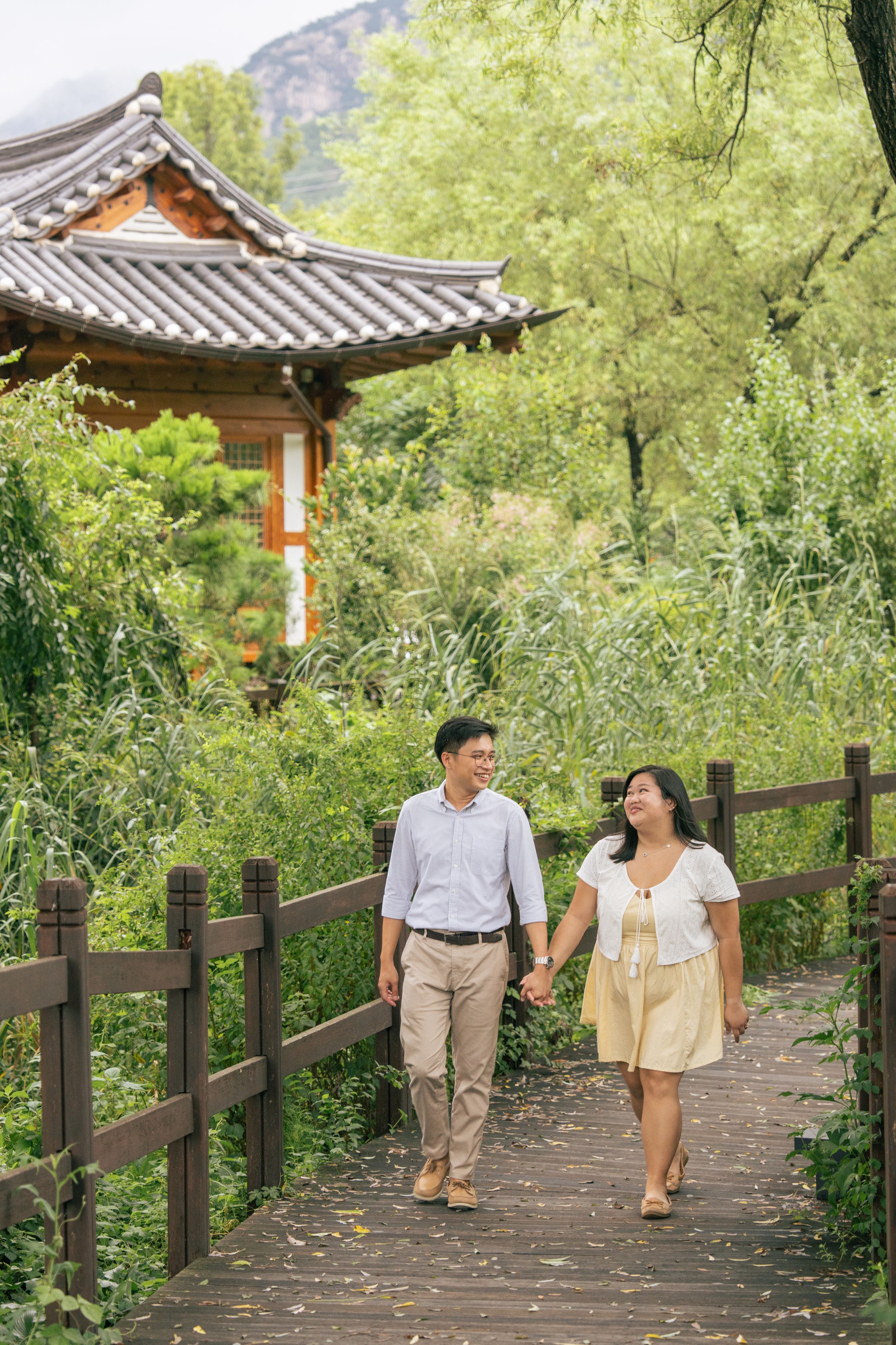 Couple walking along wooden pathway surrounded by lush greenery and traditional Korean architecture.