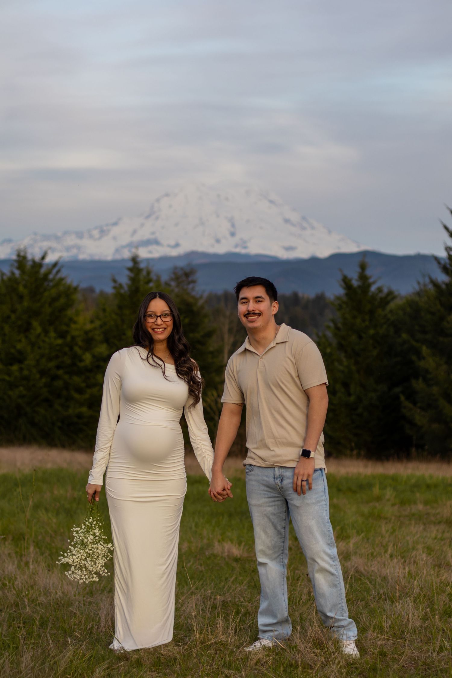 Smiling pregnant couple holding hands in field with Mount Rainier behind them taken by photographer in eatonville washington