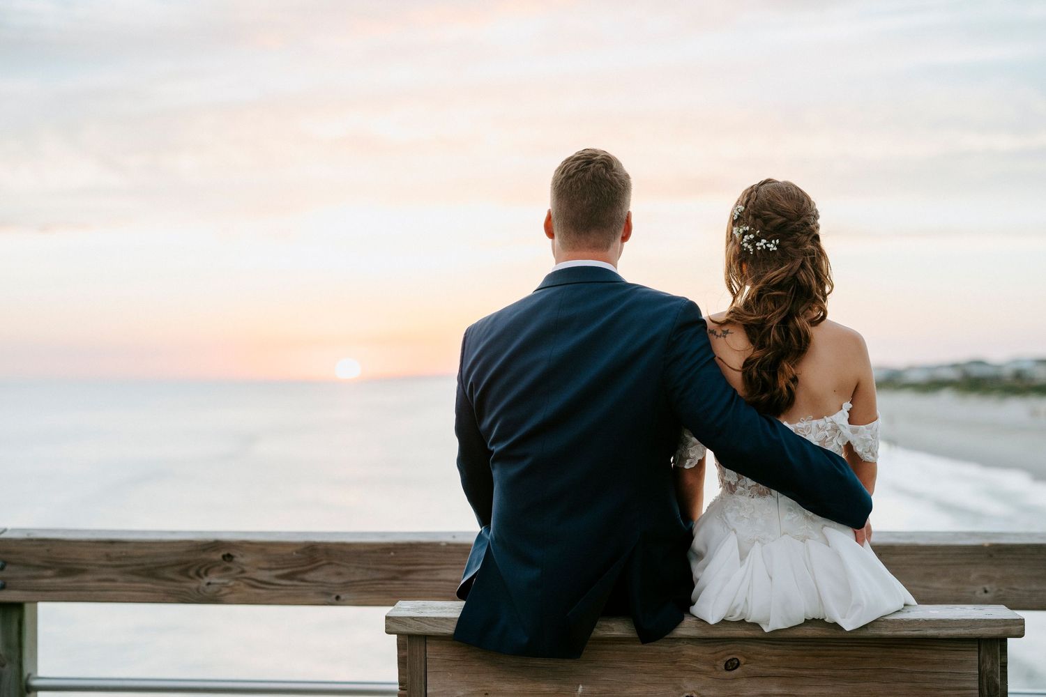 A romantic couple sits on a boardwalk overlooking the beach at sunset during their destination wedding.
