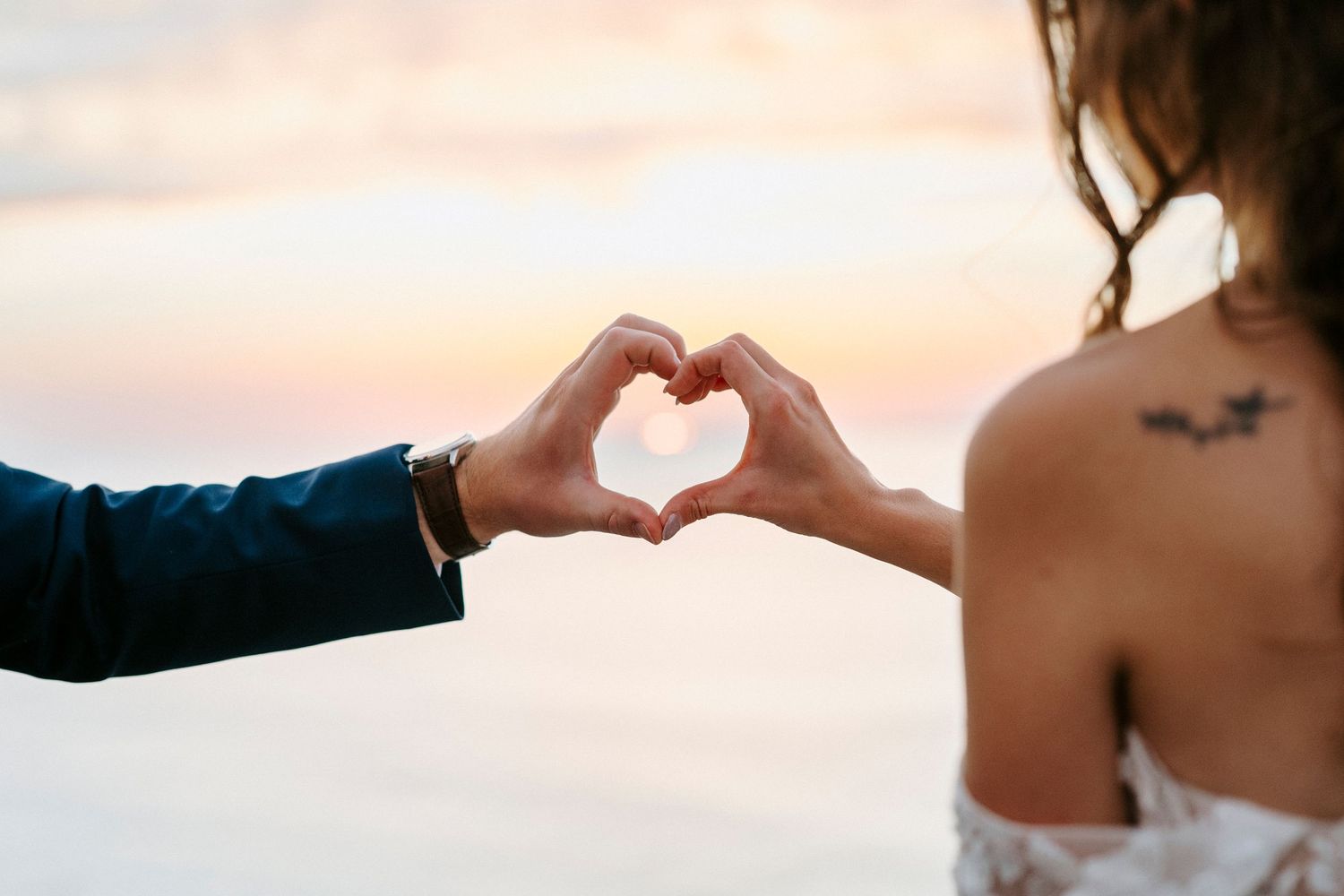 Two hands forming a heart shape gesture against a beautiful sunset sky backdrop over water.