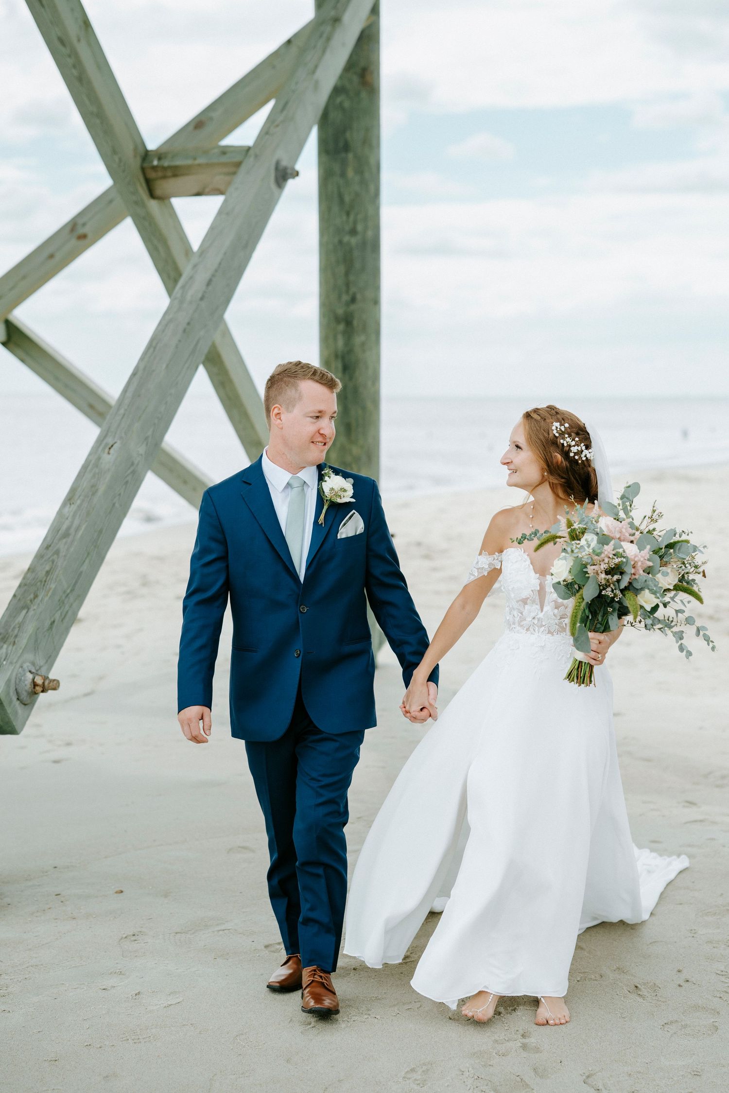 Bride and groom share intimate moments on a beach beneath weathered wooden pier supports on their wedding day.