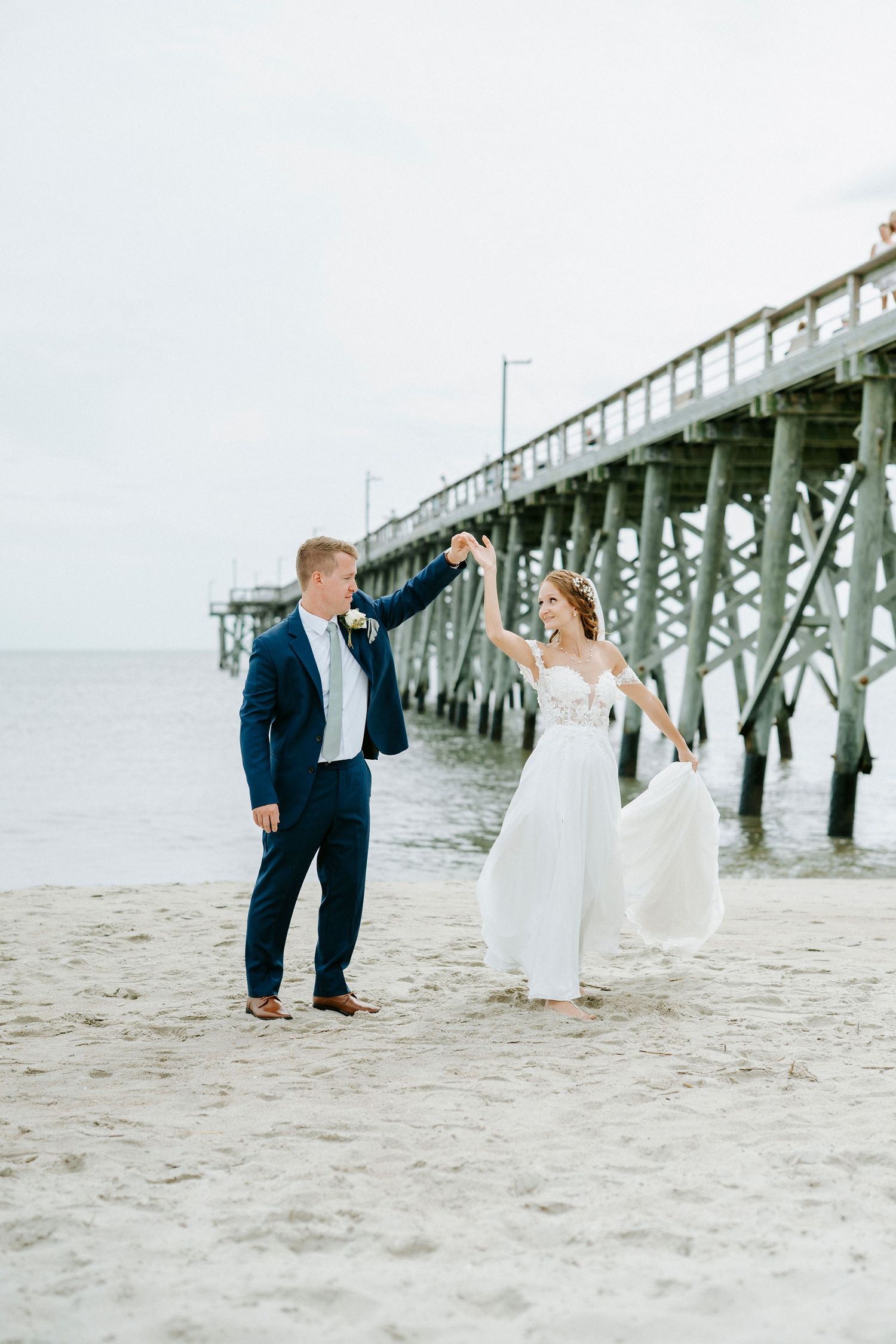 A couple celebrates on a sandy beach with a wooden pier in the background during a wedding photo session.