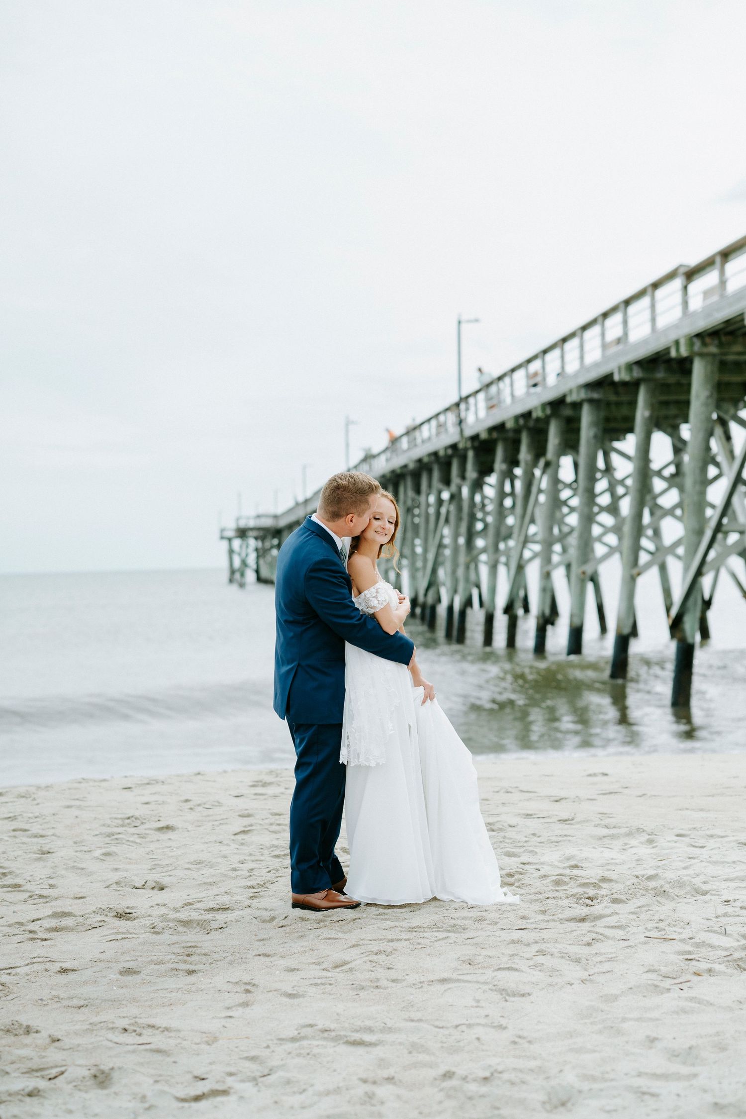 A couple shares a romantic moment on the beach near a wooden pier during their seaside wedding photoshoot.