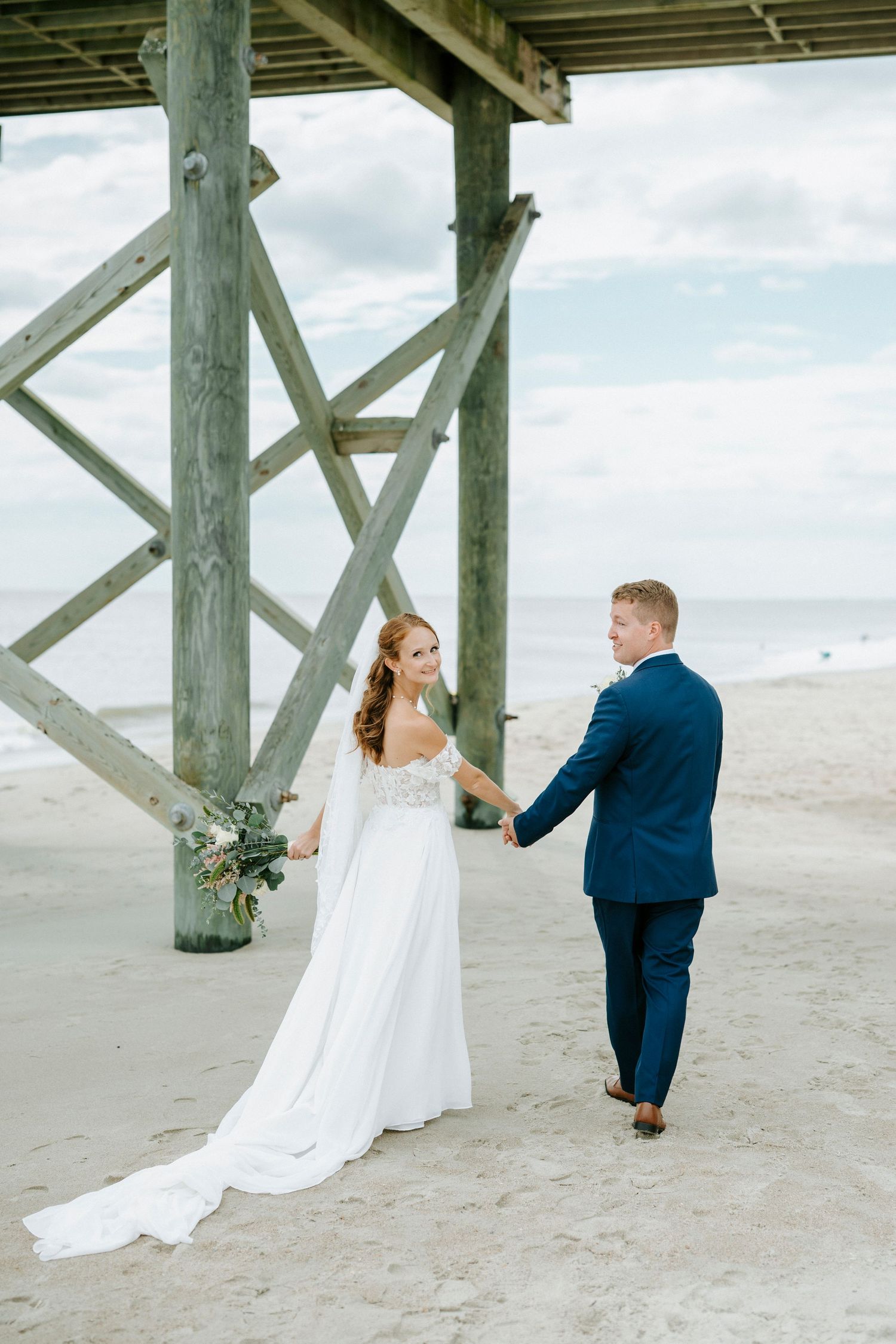 A couple poses playfully under a wooden pier during their beach wedding photo session.