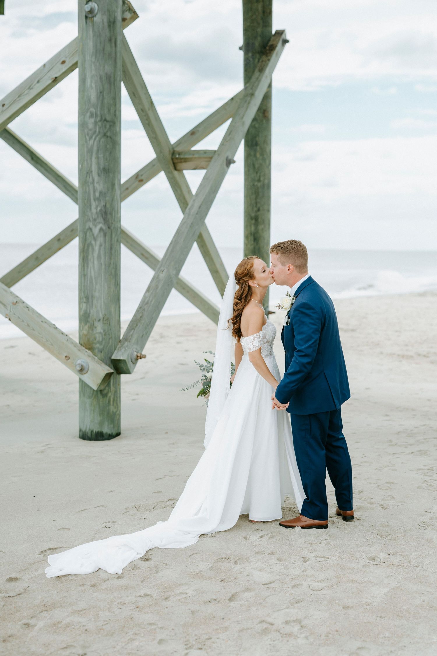 A romantic beach wedding photograph series showing a couple by wooden pier posts on a cloudy day.