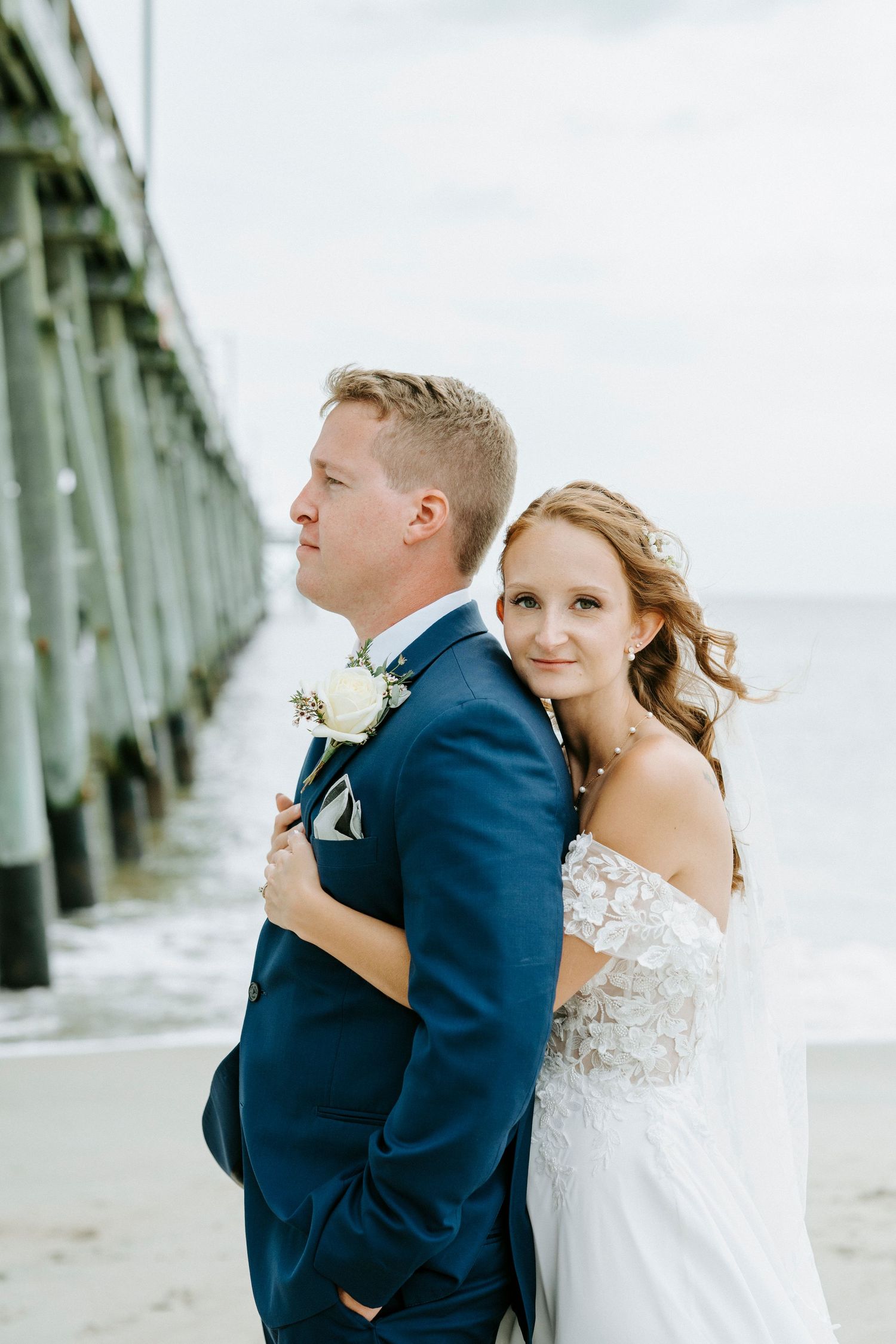 Wedding couple embraces on a sandy beach with a long wooden pier in the background.