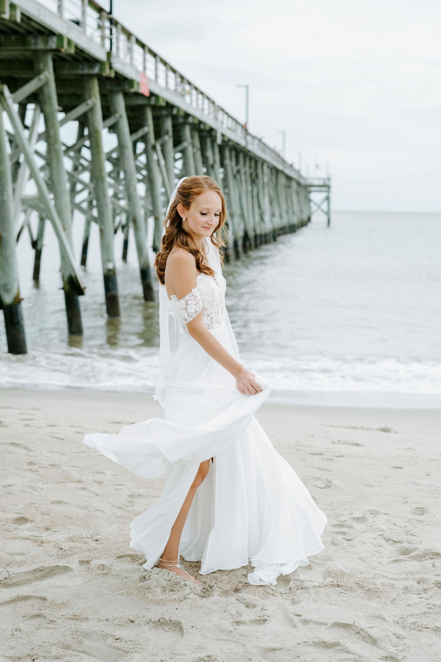 A bride in white dress twirls joyfully on the beach near a coastal pier.