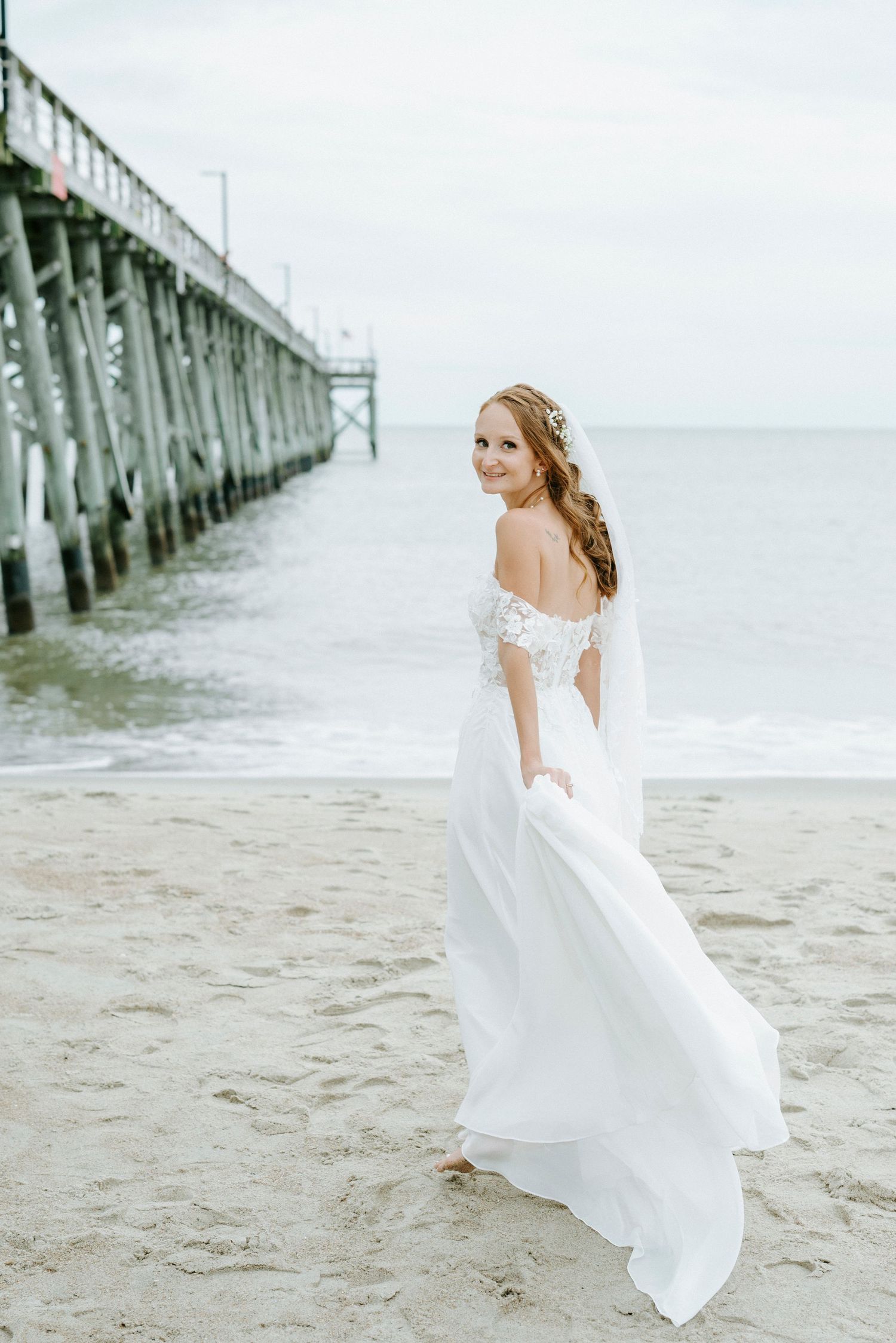 A flowing white wedding dress photographed at a beach pier in soft natural lighting.