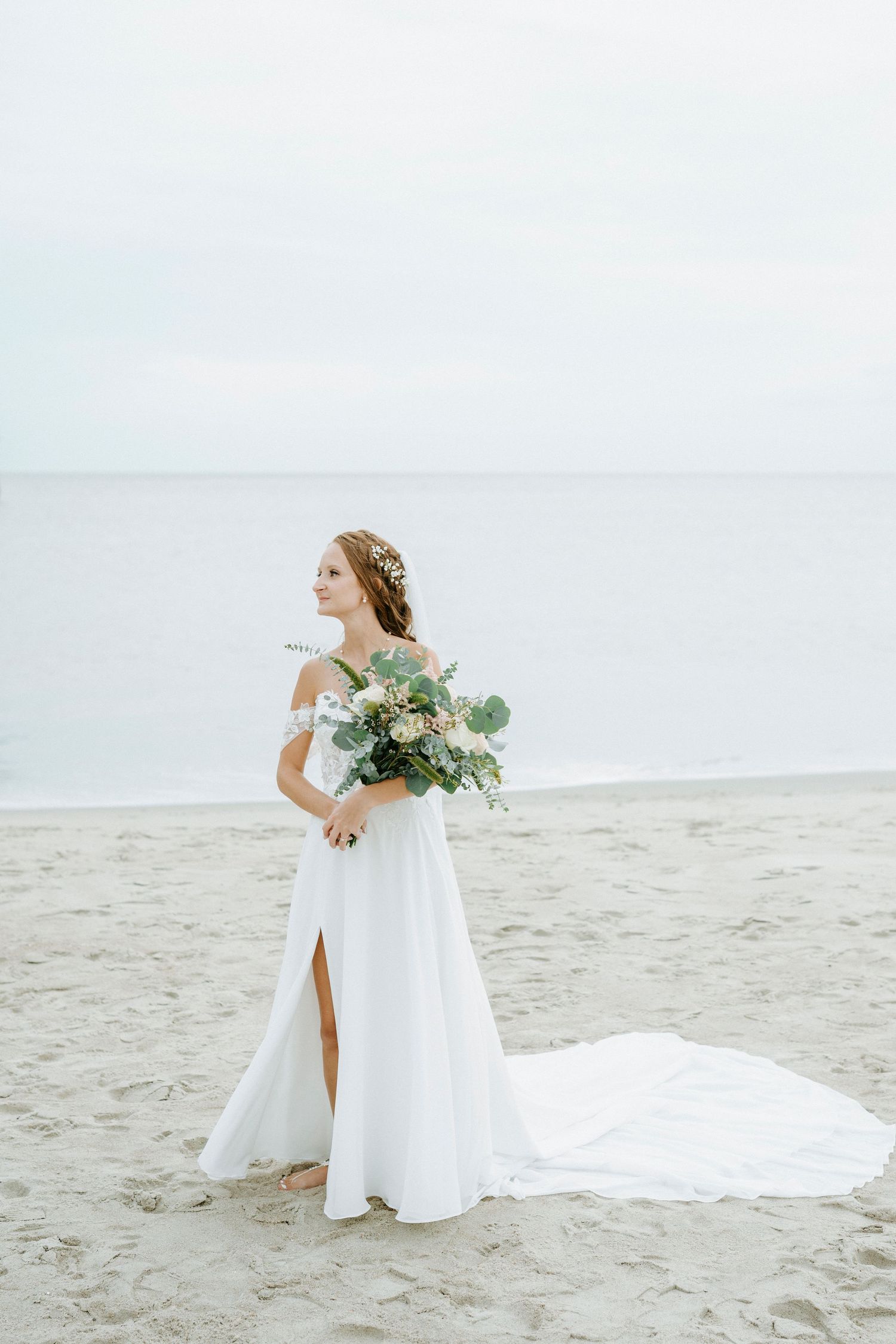 Bride with flowing white dress and bouquet poses gracefully on sandy beach near ocean pier.
