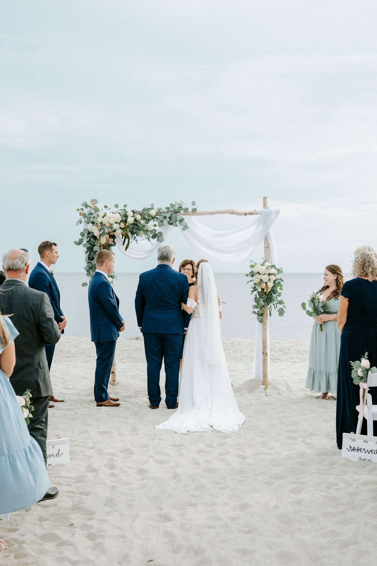 Beachside wedding ceremony setup with white arch and floral decorations as guests gather on sandy shore.