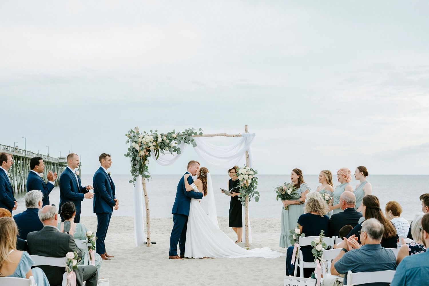 A beach wedding ceremony unfolds with white chairs and floral arrangements against an ocean backdrop.