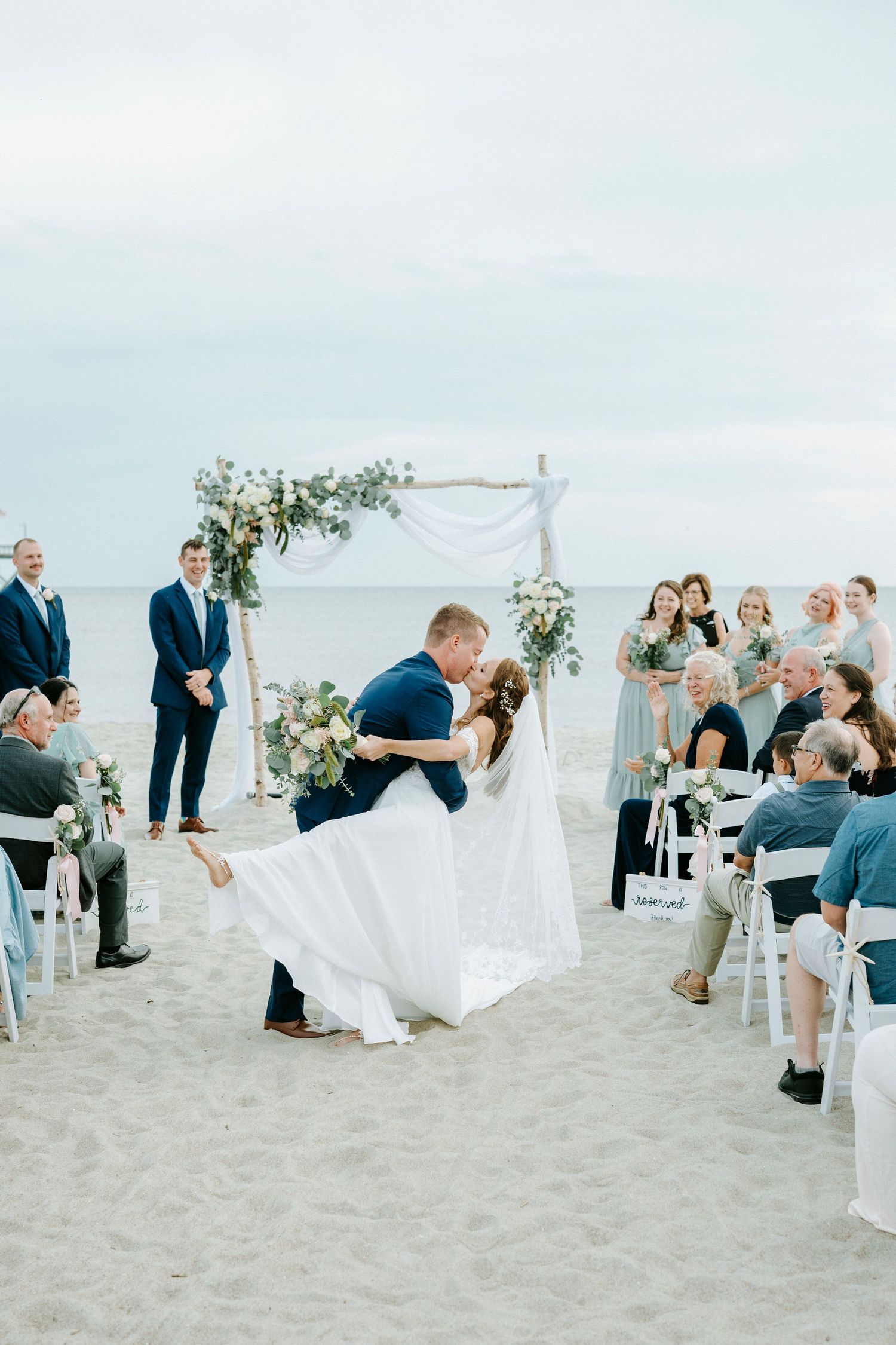 Beachside wedding ceremony setup with white chairs and floral arbor on sandy shores.