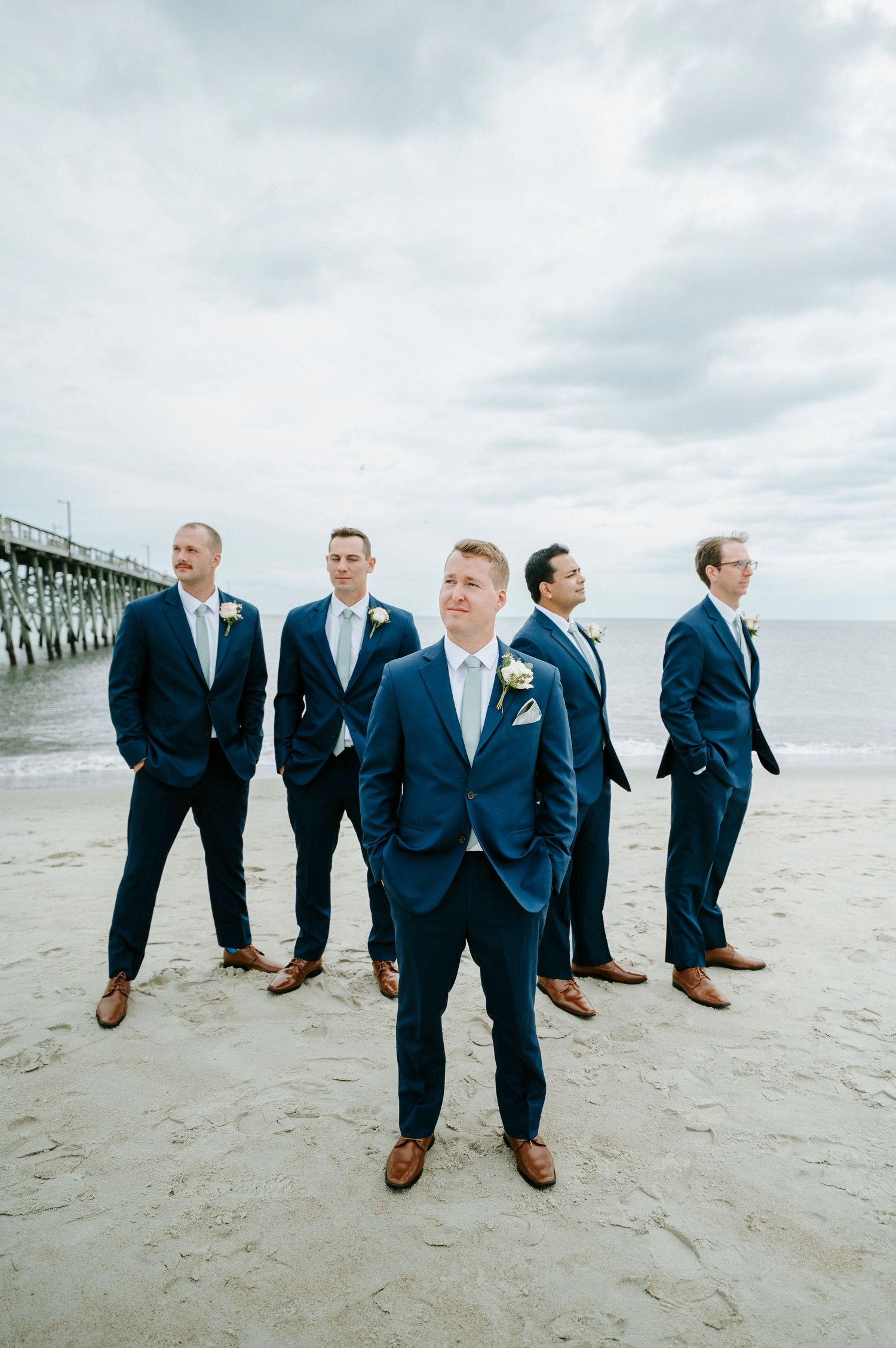 A group in navy suits and brown shoes pose playfully on a beach with a pier in the background.