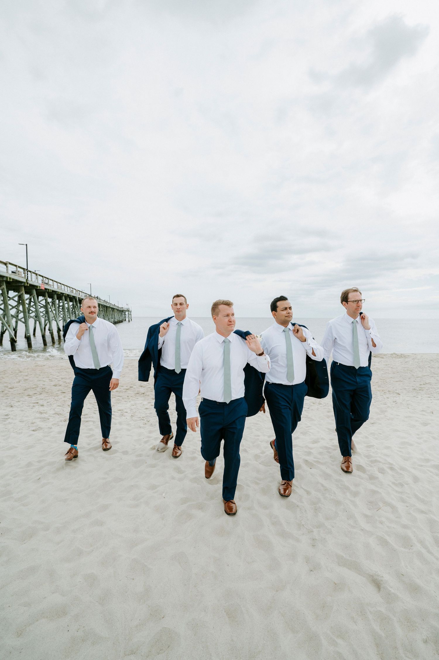 Wedding party wearing white shirts and navy pants jump together on a sandy beach near a pier.