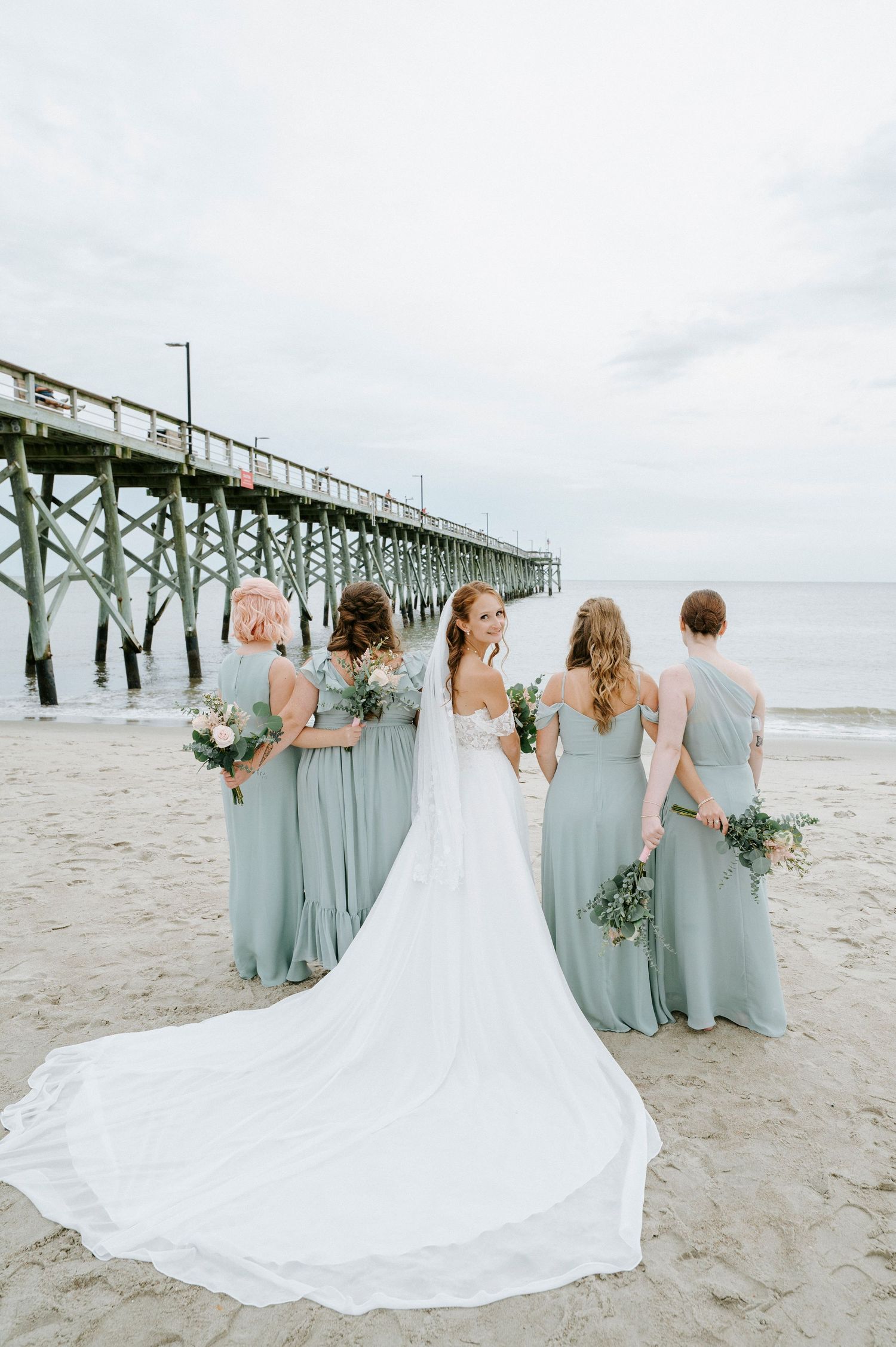 A group in elegant blue bridesmaids dresses and a flowing white wedding gown pose together on a beach near a pier.