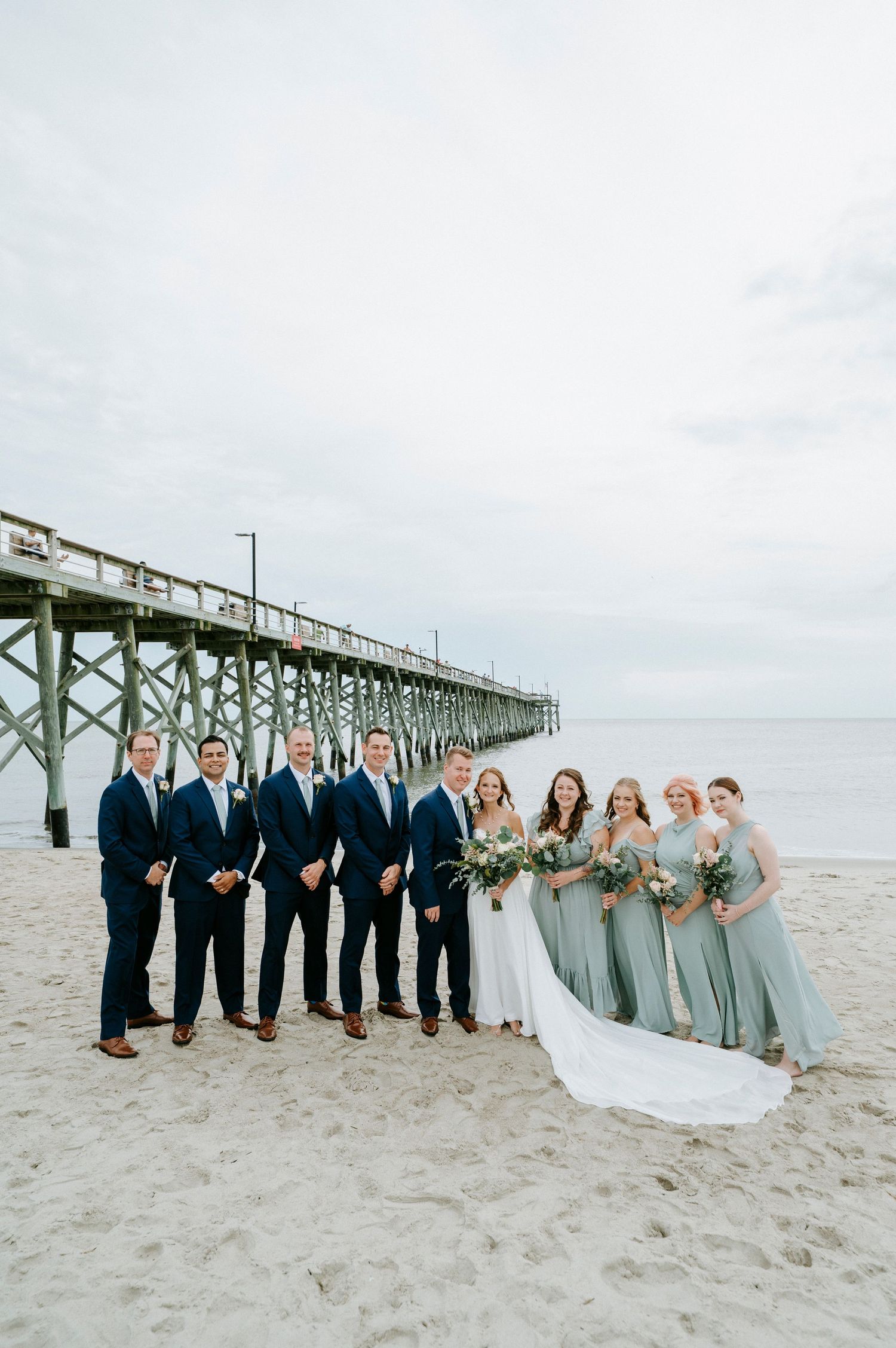 Wedding party in navy and gray attire poses together on beach with pier backdrop.