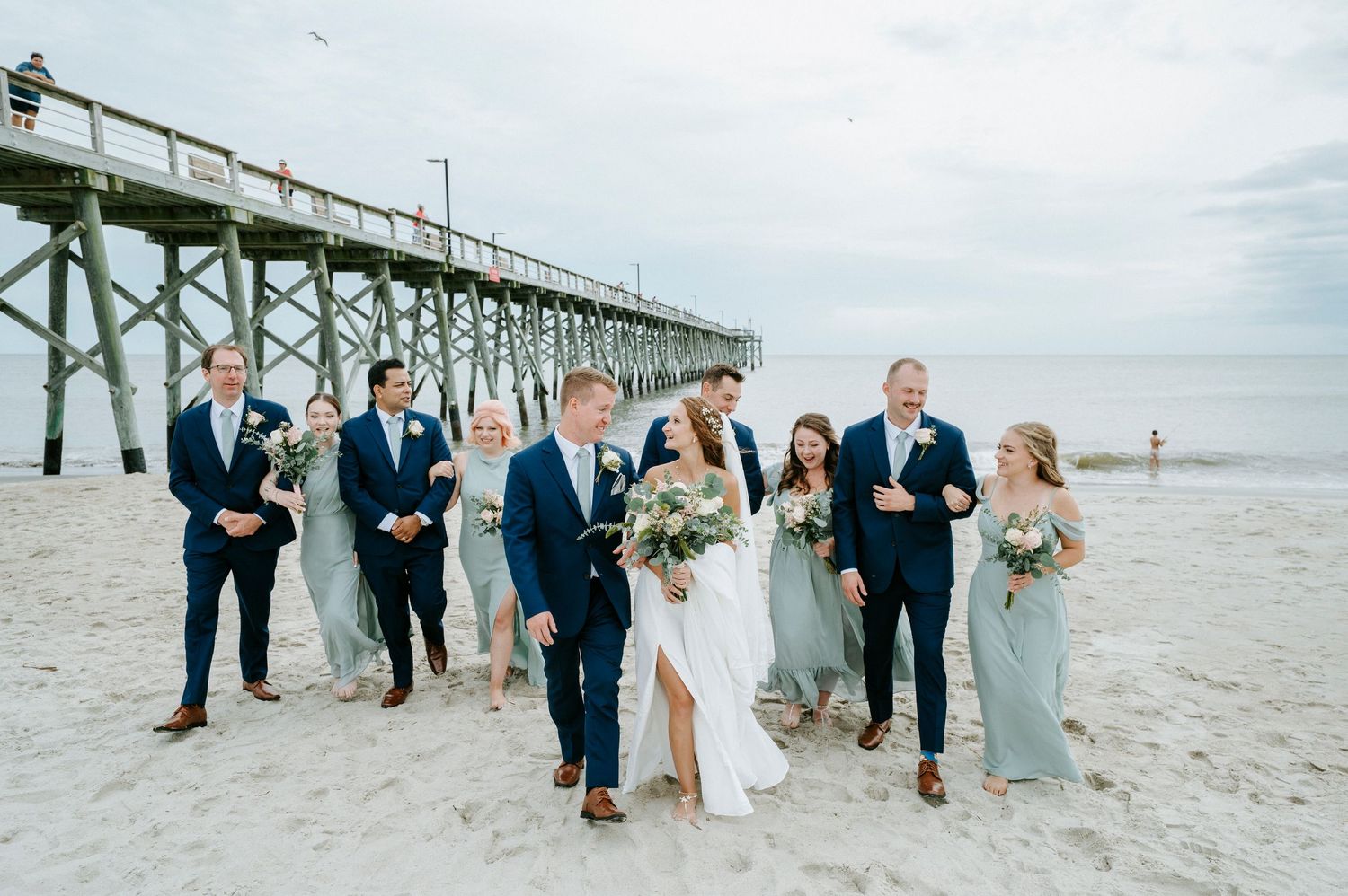 Large wedding party lined up for formal photos on beach with wooden pier in background.