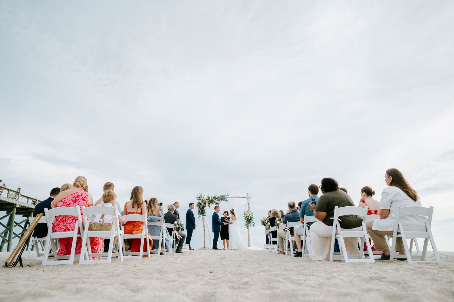 Beach wedding ceremony setup with white chairs and guests along shoreline.