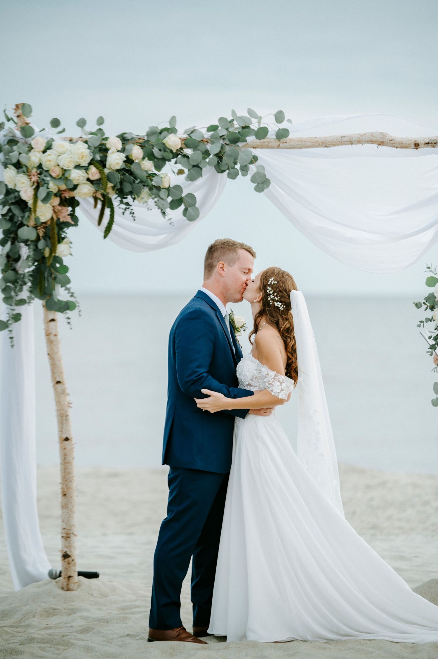 Beach wedding ceremony under a birch wood arch decorated with eucalyptus and white fabric.