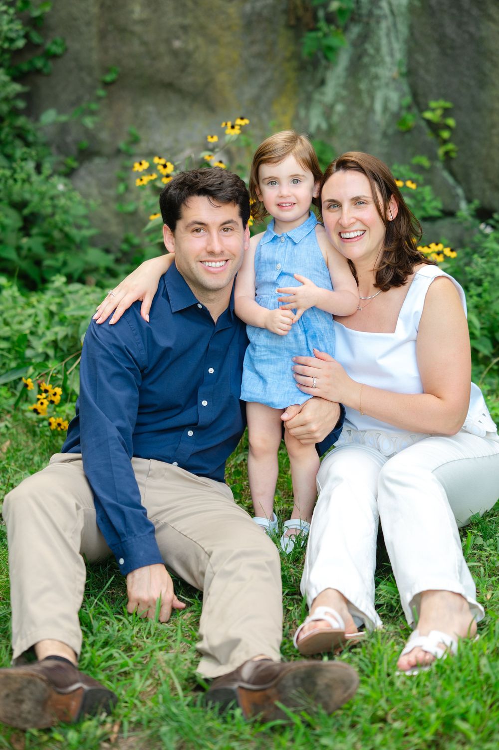 A family sits together on grassy ground in Central Park with yellow flowers blooming nearby.