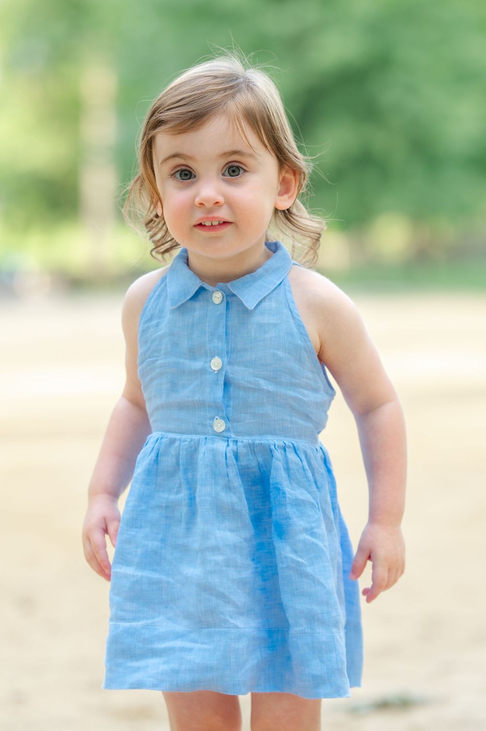 A toddler in a sleeveless light blue dress stands outdoors with a blurred green background in Central Park during a family photo session.