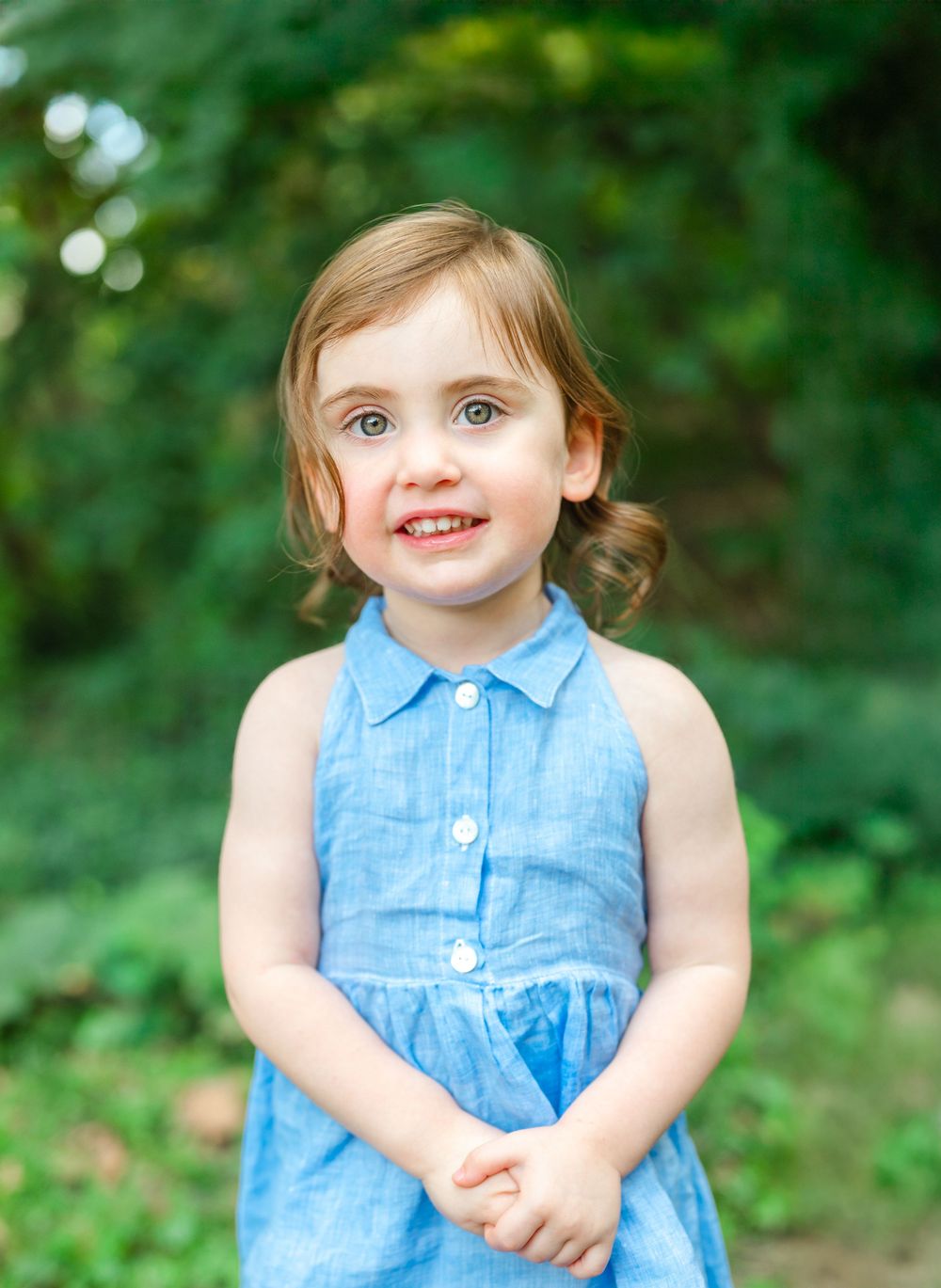A small child wearing a sleeveless blue dress poses outdoors against a green natural background.