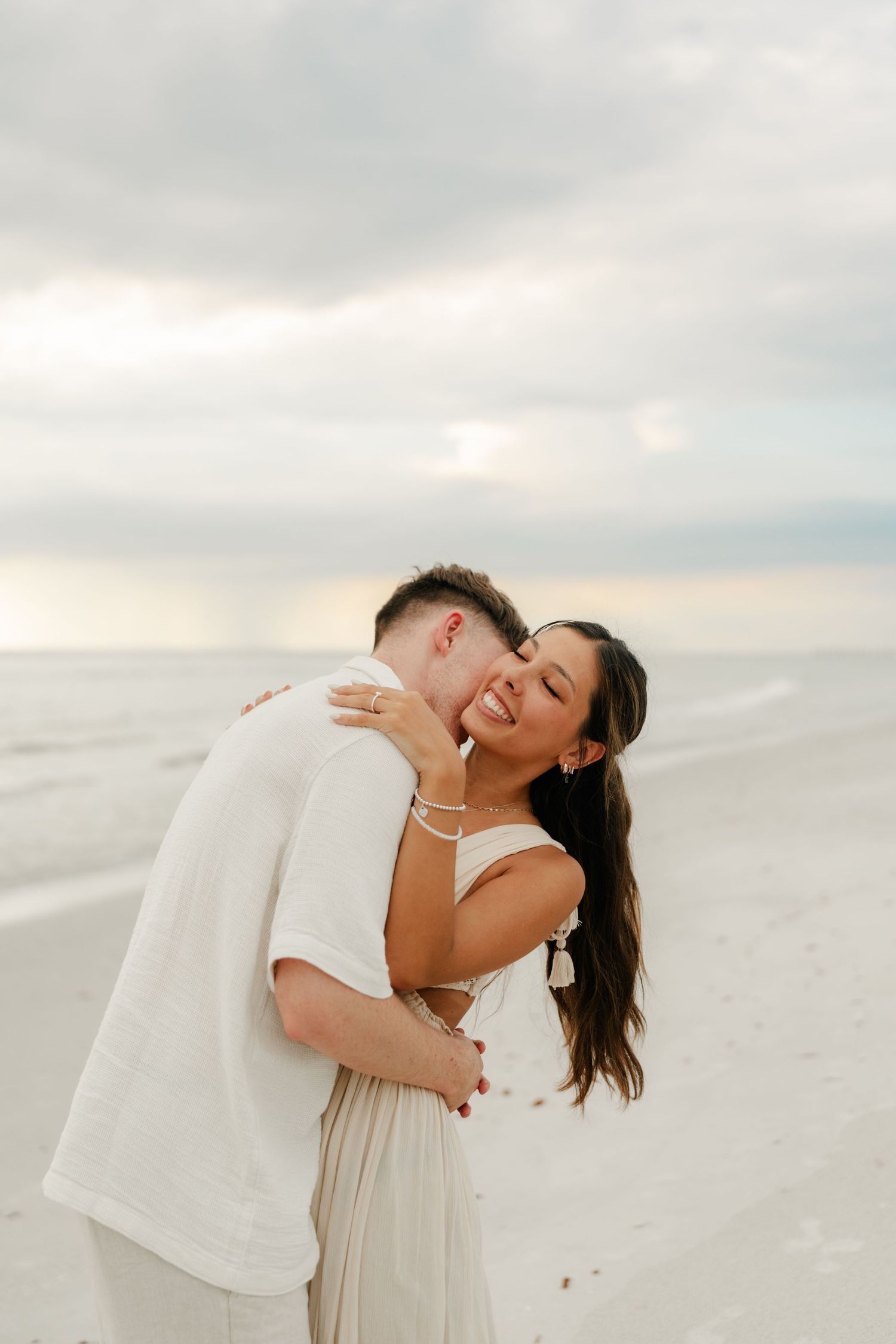 A couple in white outfits embraces joyfully on a cloudy beach in a romantic photo series.