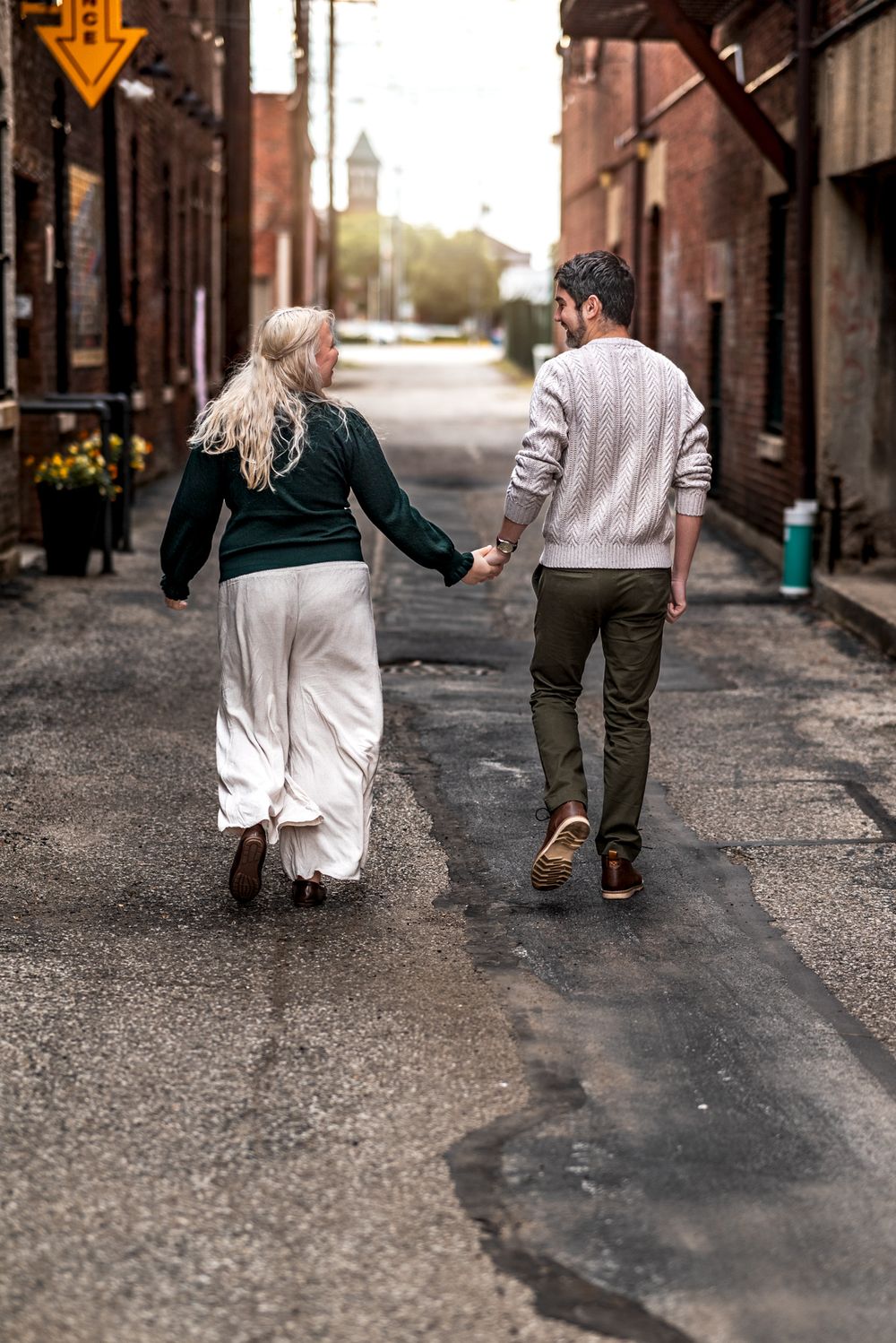 A couple walks hand in hand down a charming brick alley with sunlit buildings and flowers in the background.