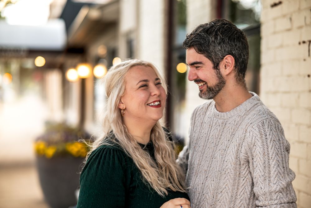 A couple shares a joyful moment together outdoors while wearing cozy sweaters against a brick wall background.