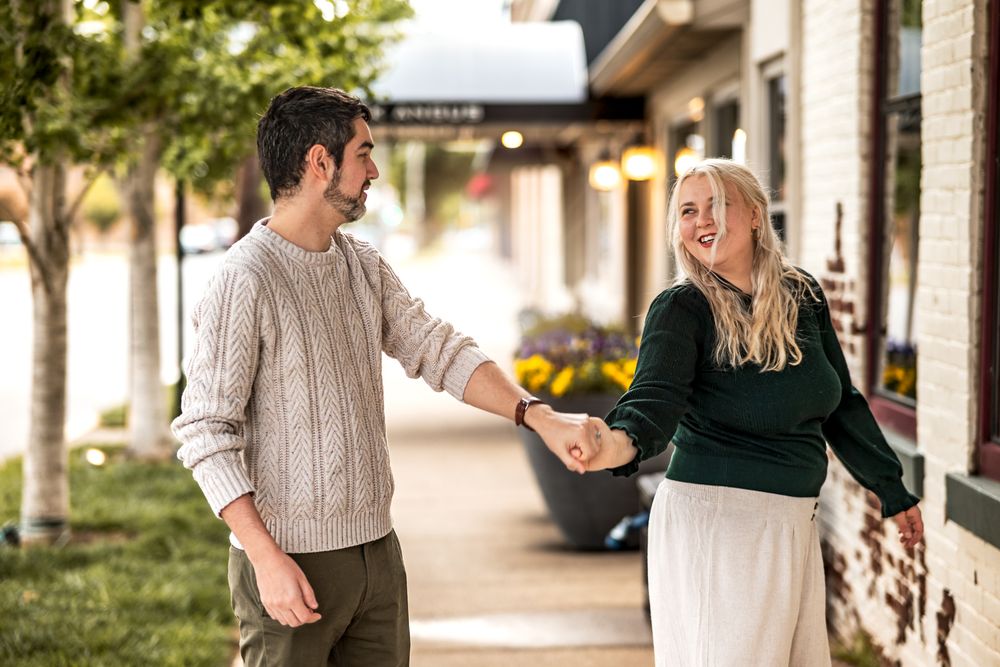 Couple walking together and laughing on a sunny sidewalk with storefronts in the background.