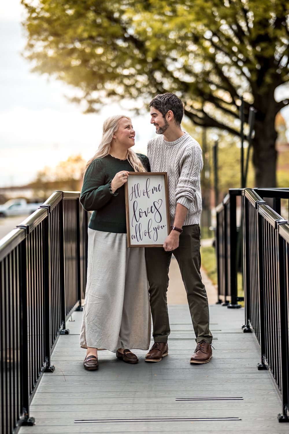 A couple sits playfully on a wooden bridge while holding signs during an autumn photoshoot.